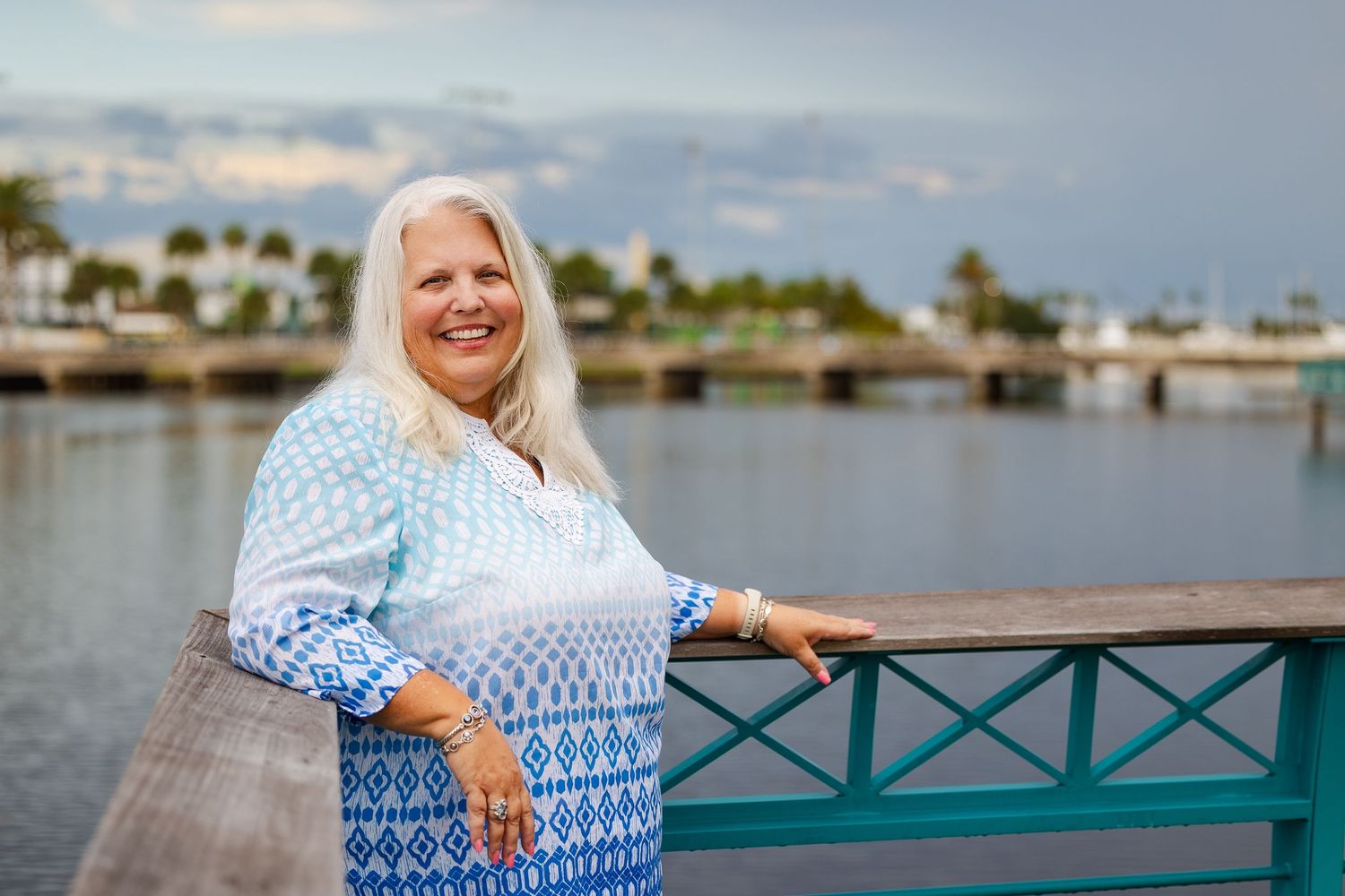 Smiling woman with long white hair leans on teal railing wearing blue patterned top by waterfront at dusk.