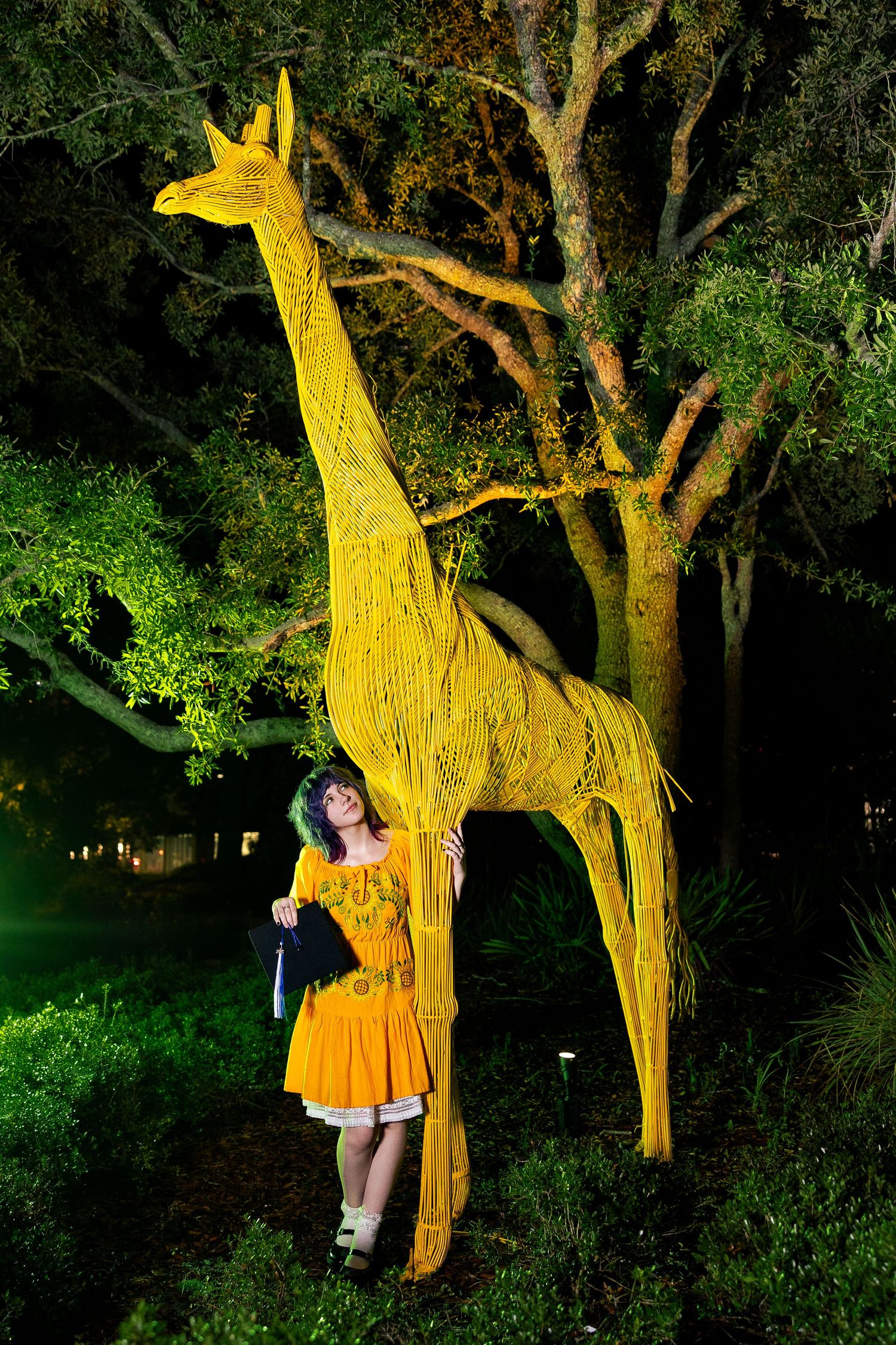 Woman in orange dress poses beside a tall yellow giraffe sculpture illuminated at night in a lush garden.