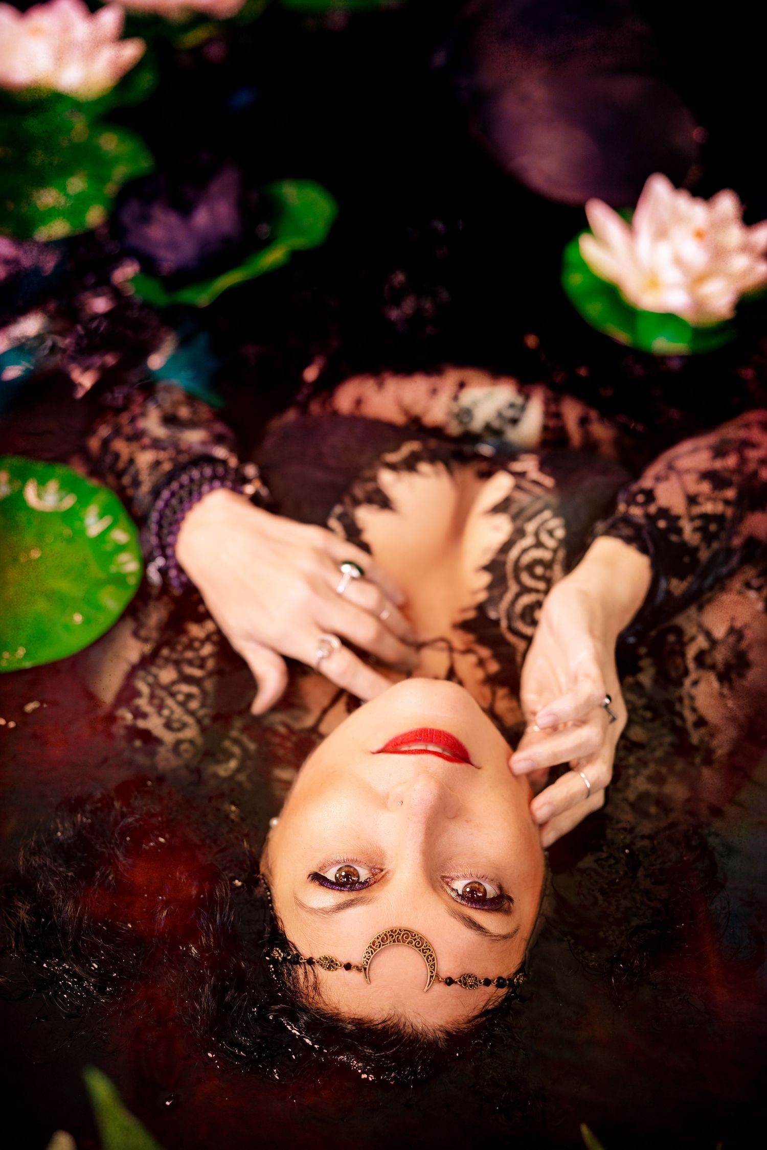Woman in black lace dress floating in dark water surrounded by lily pads and pink lotus flowers.