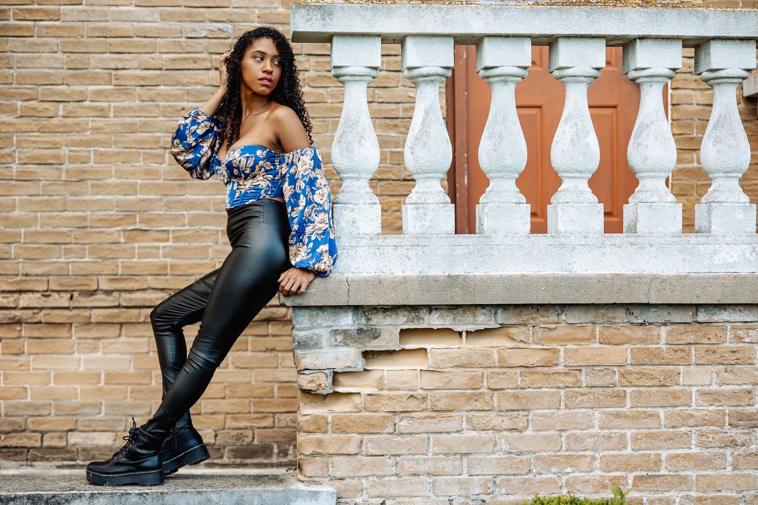 Woman in blue floral off-shoulder top and black leather pants poses by ornate stone balustrade.