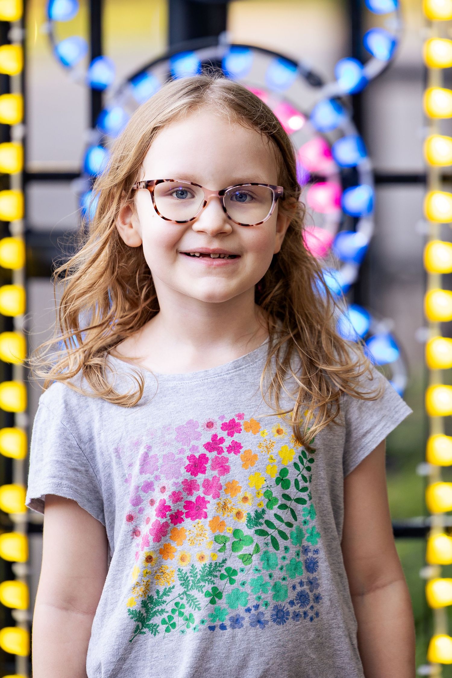 Smiling young girl with glasses wearing a colorful floral t-shirt stands in front of bright lights.