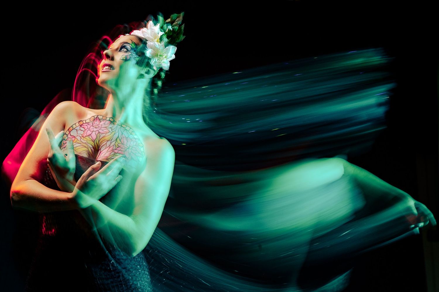 Long exposure photo of dancer in green light with white flower crown against black background.