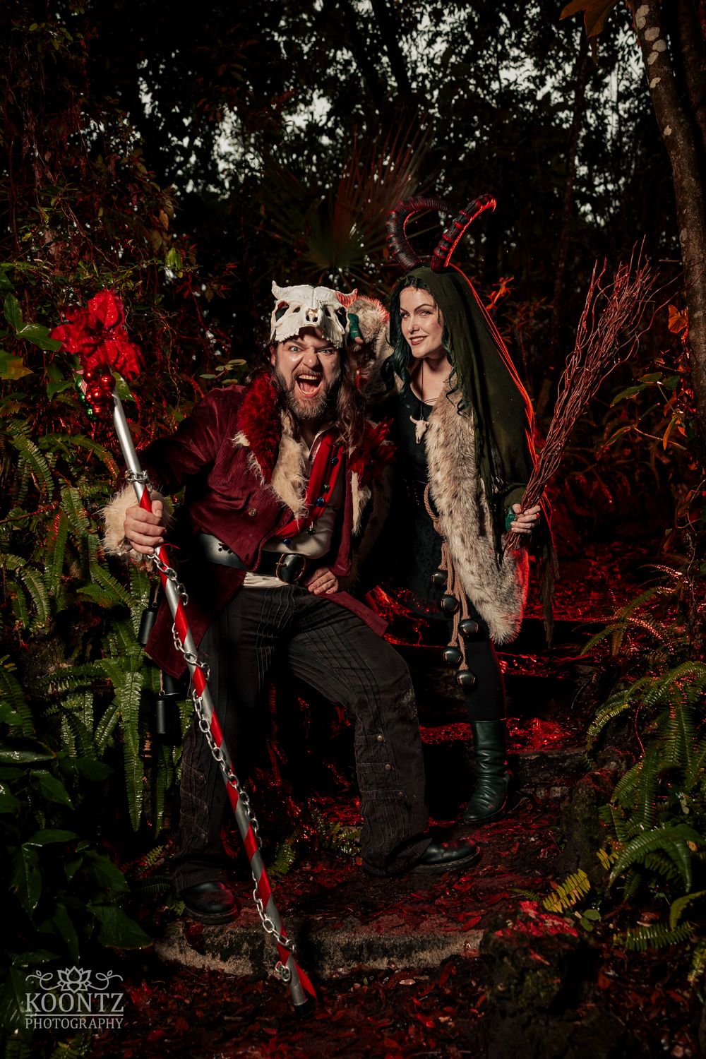 Couple in elaborate fantasy Halloween costumes pose with staff and broom in dramatic red-lit forest setting.