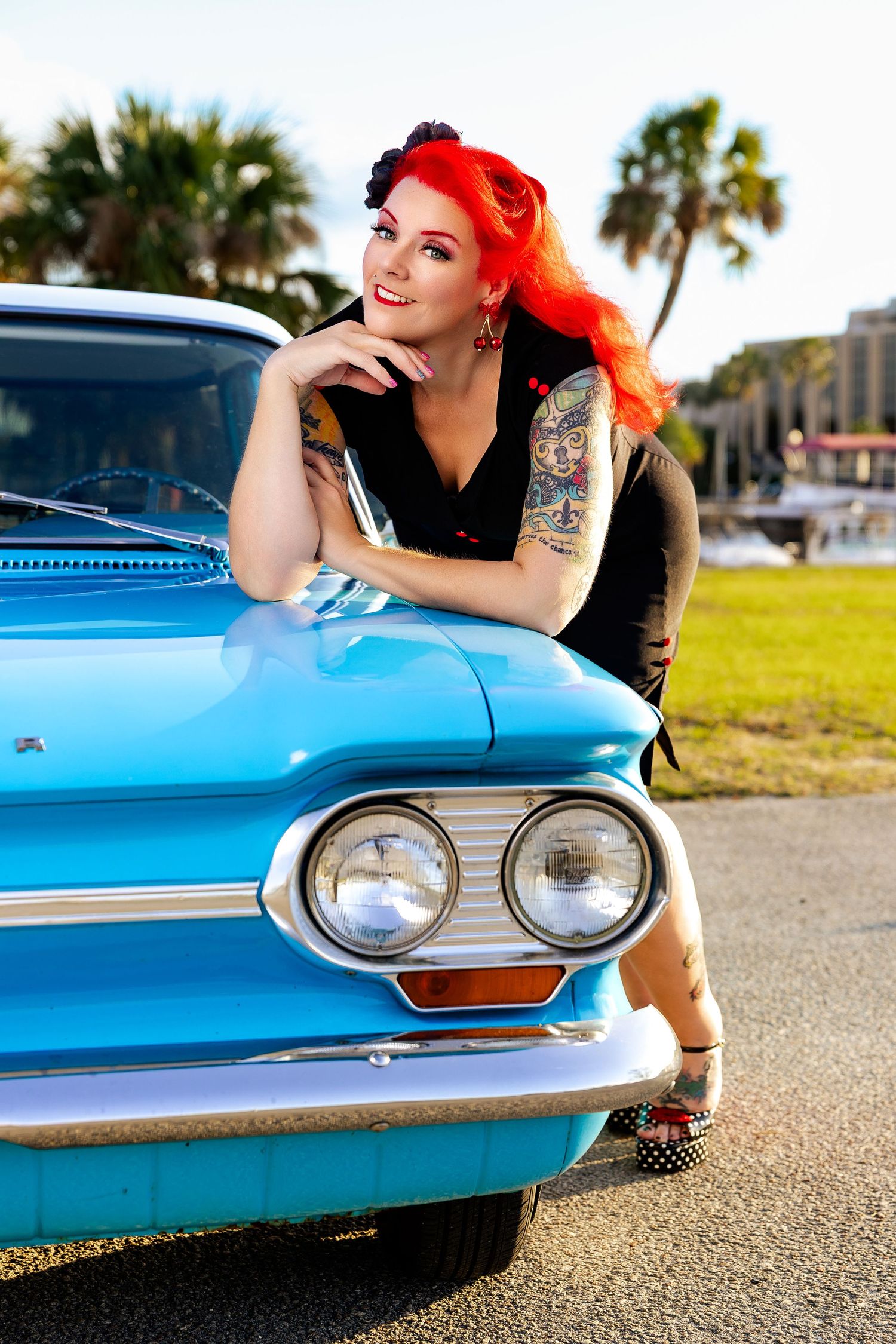 Smiling tattooed redhead pinup woman leaning on front of blue vintage classic car.