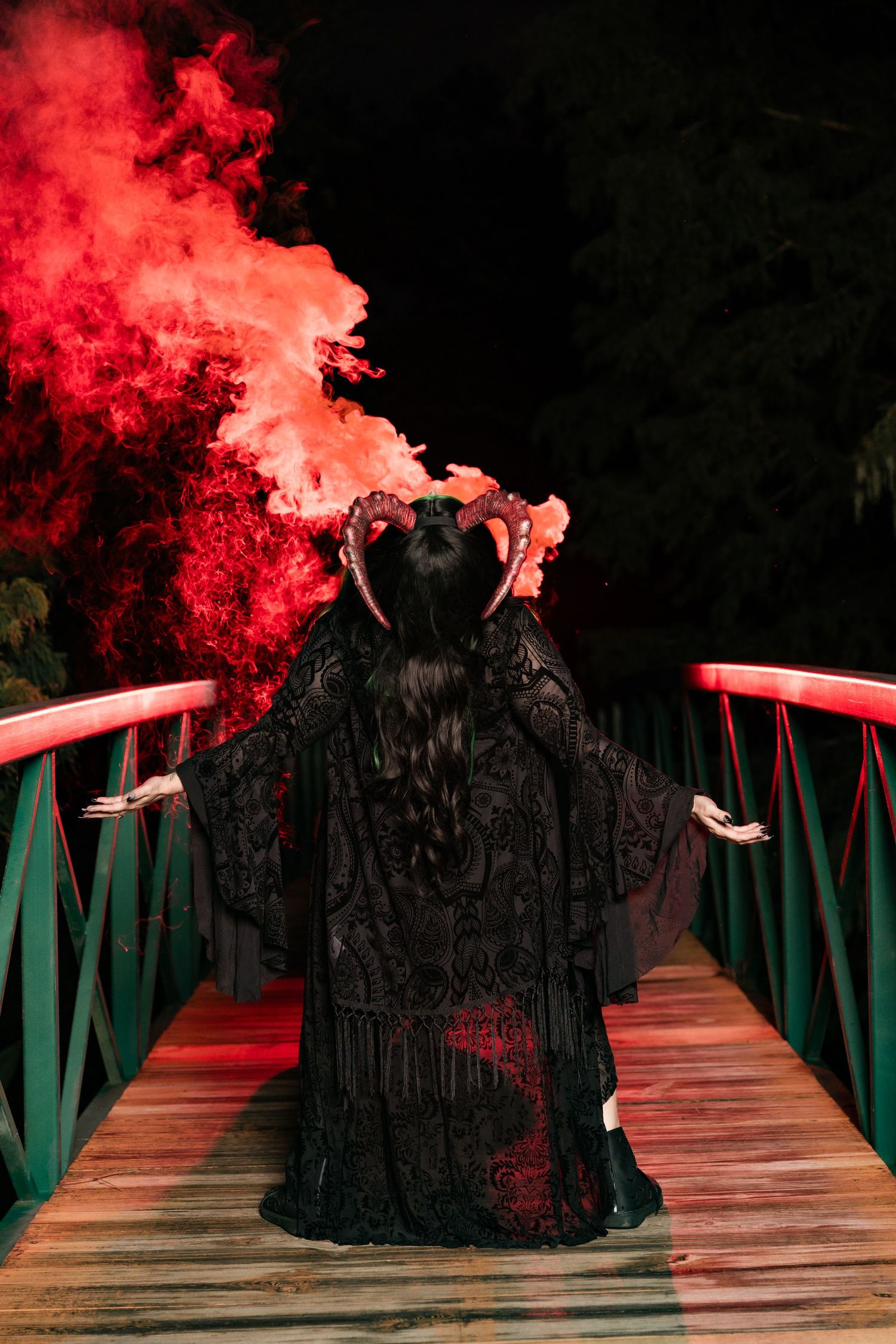 Woman in ornate black gown with horned headpiece stands on a bridge surrounded by dramatic red smoke at night.
