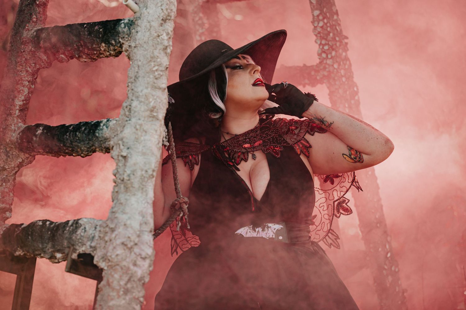 Woman in black witch hat and red lace dress poses dramatically in red smoke outdoors.