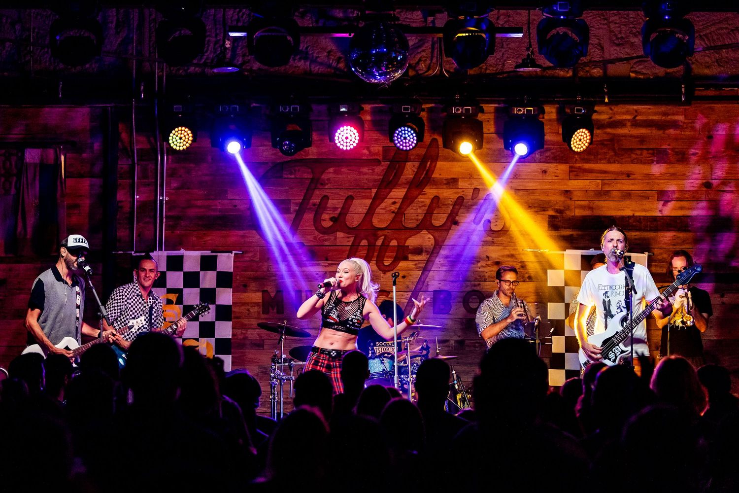Live band performing on colorfully lit stage with checkered backdrop at indoor music venue.