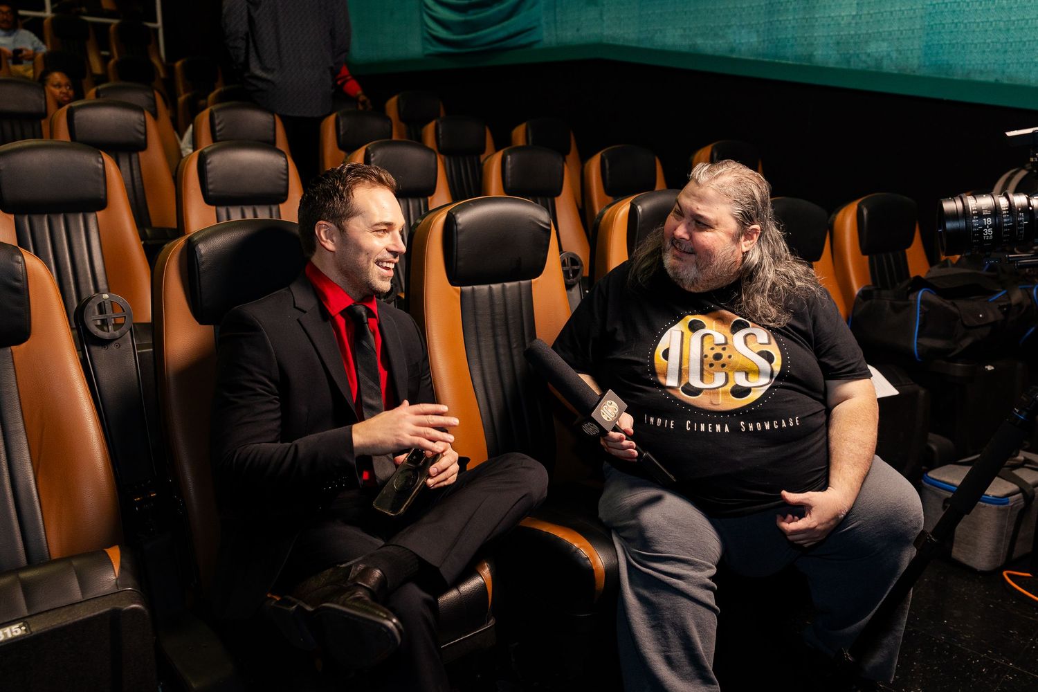 Two men in formal attire sit and converse in luxury cinema seating at a movie premiere.