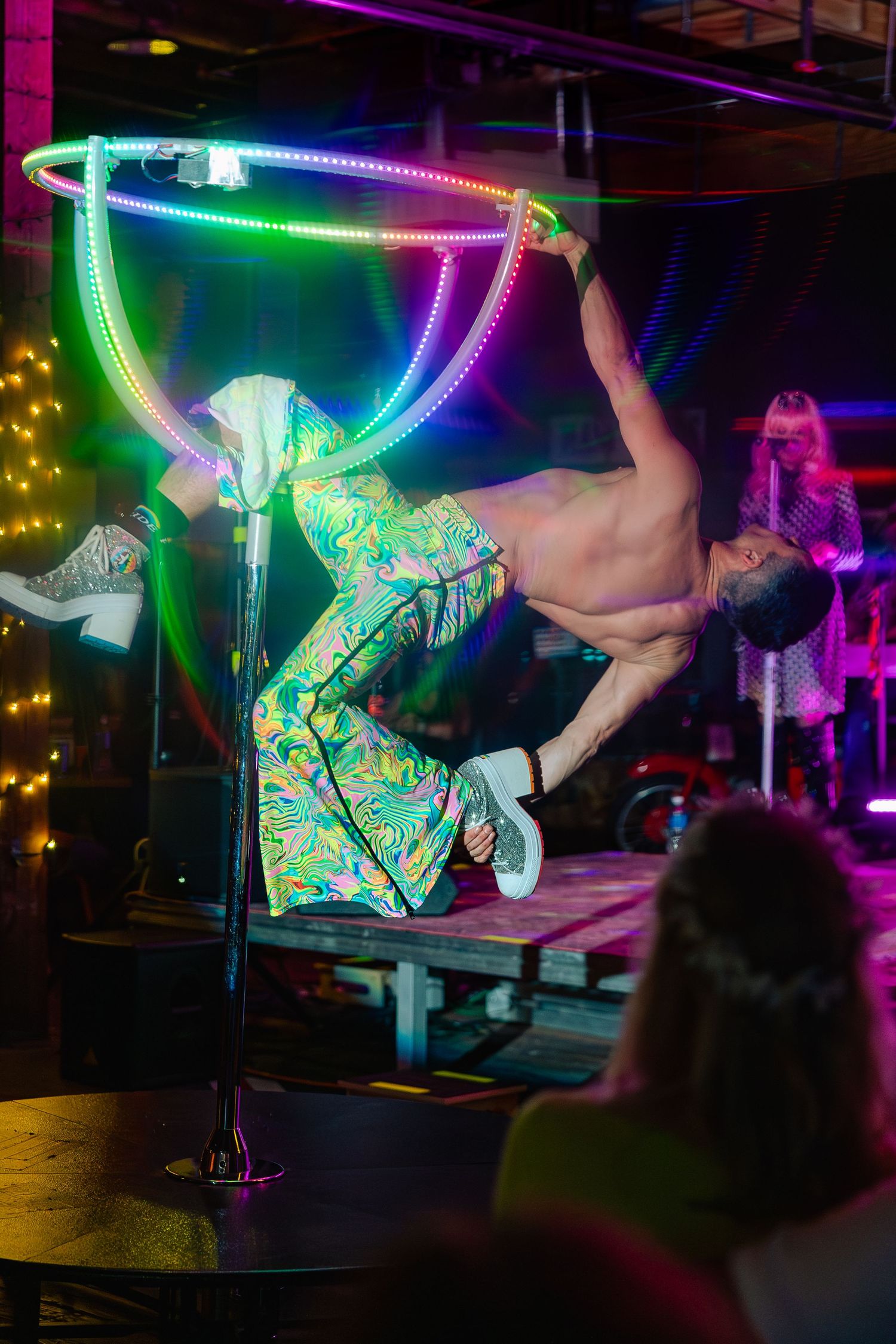 Acrobat performing aerial hoop act under rainbow LED lights on stage at a colorful nightclub event.