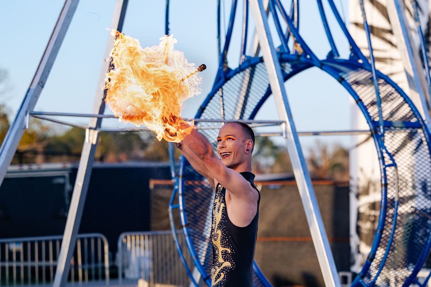 Smiling fire performer holding blazing fire prop beside a large metal gyroscope at an outdoor event.