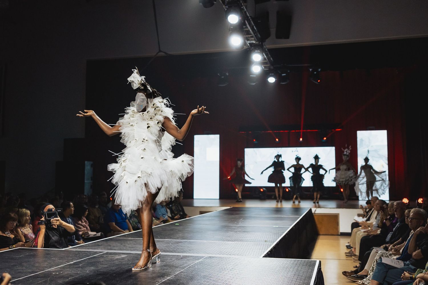Model in white feathered mini dress and headpiece struts runway at avant-garde fashion show.