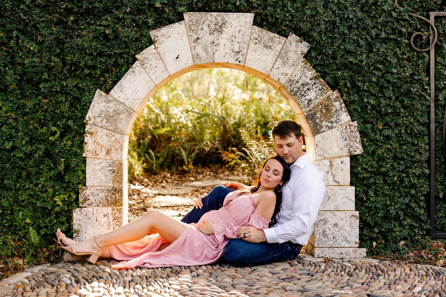 Couple relaxing together in a stone archway surrounded by ivy, woman in pink maternity dress, man in white shirt.