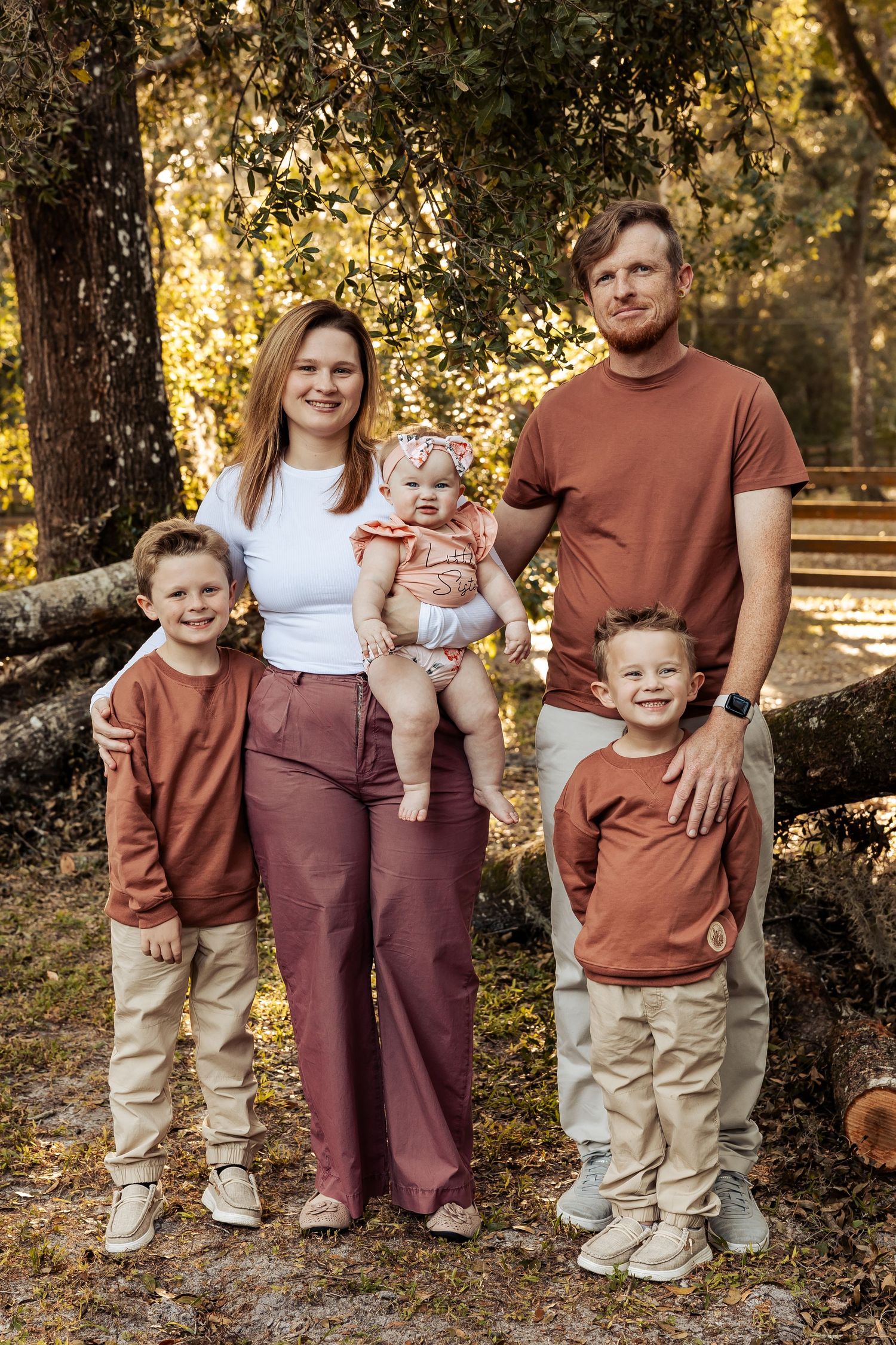 Family of five standing together in a sunlit forest, wearing coordinating rust, white, and tan outfits.