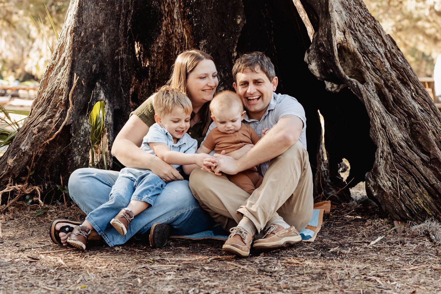 Joyful family of four sitting together at the base of a large tree outdoors.