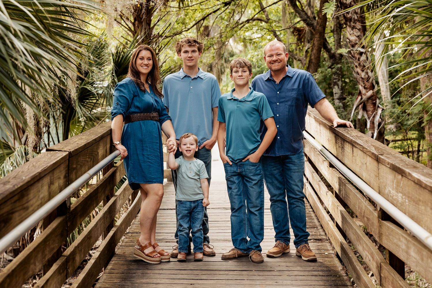 Family of five in coordinating blue outfits posing on a wooden bridge surrounded by tropical greenery.