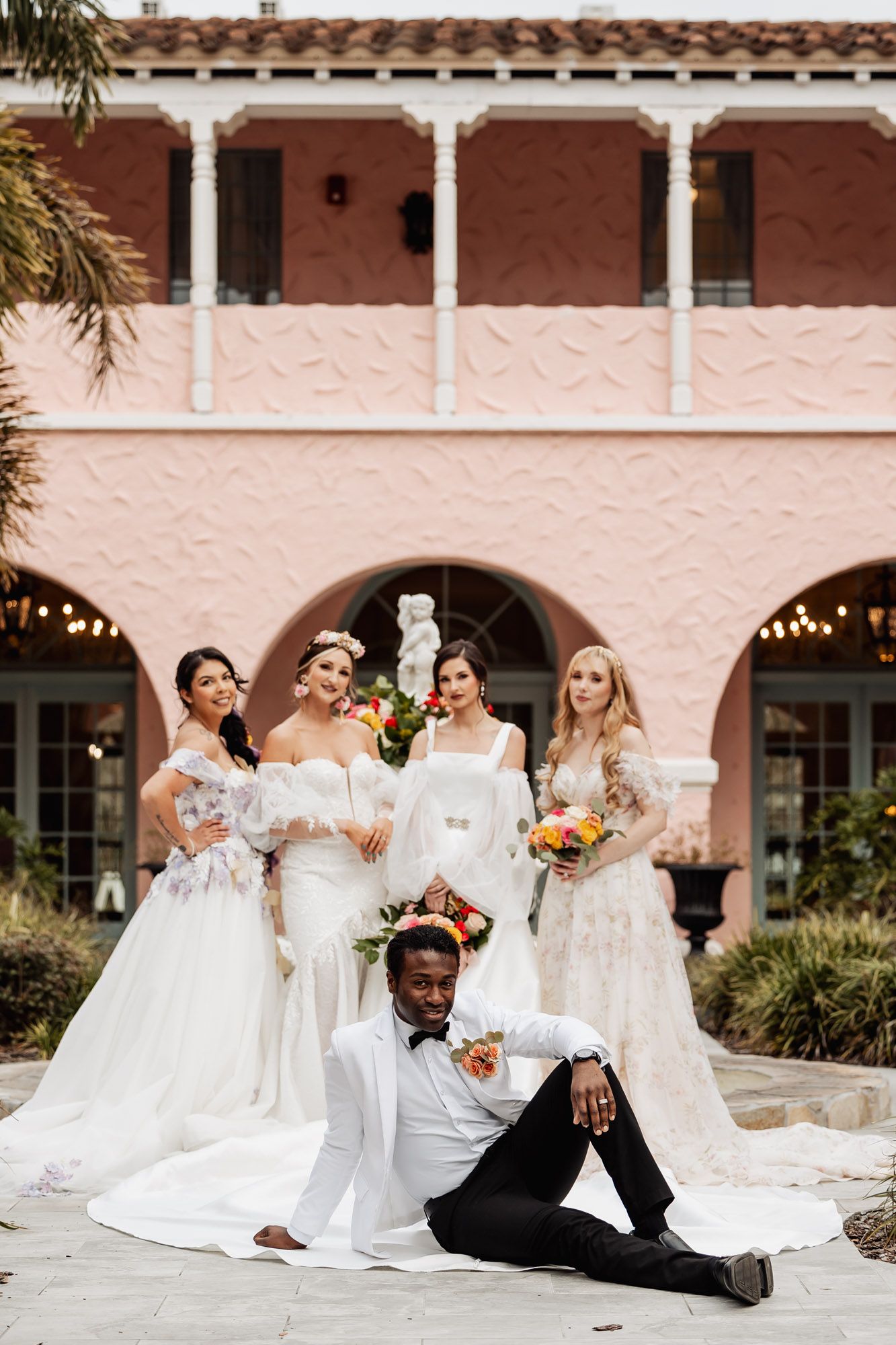 Four brides in white gowns pose with a groom in a white suit outside a pink venue.