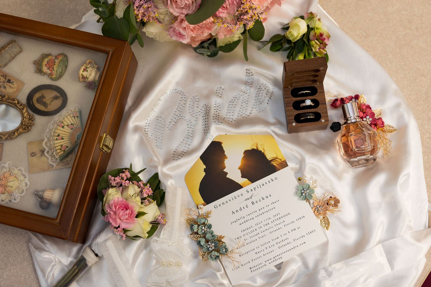 Wedding flat lay featuring invitation, floral bouquet, vintage jewelry, and perfume on white satin.