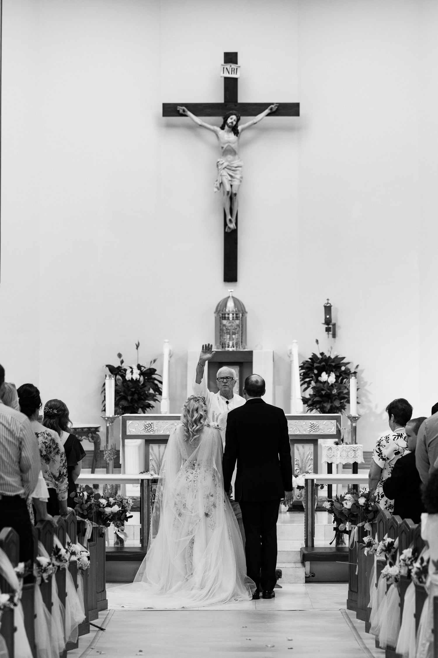 Black and white photo of bride and groom standing at church altar beneath large crucifix.
