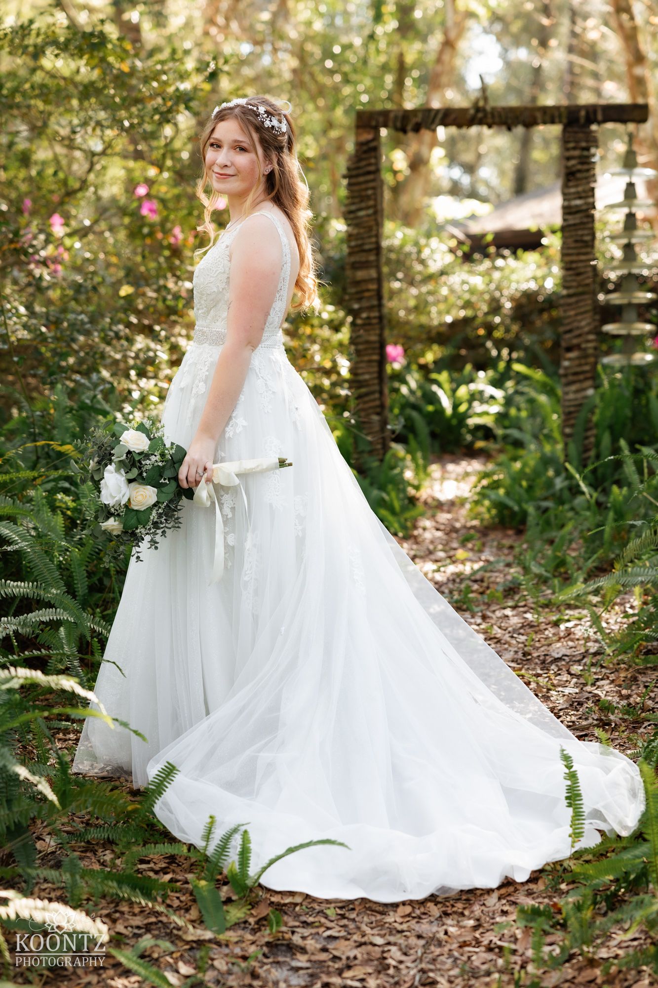 Bride in a white lace gown holds a white rose bouquet in a lush garden wedding setting.