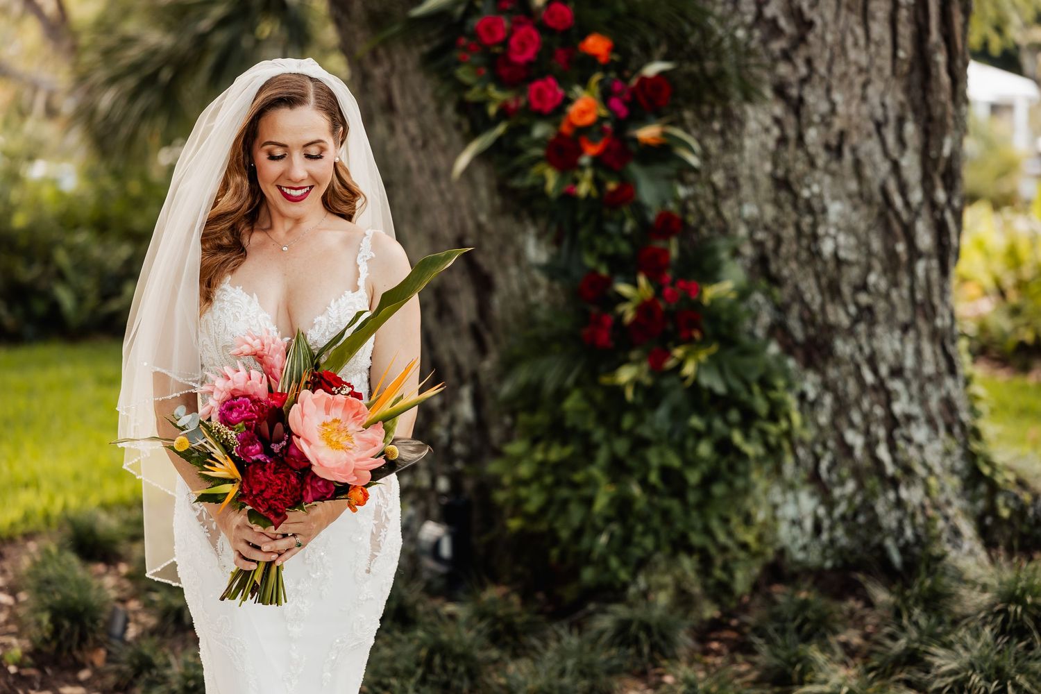 Smiling bride in lace wedding dress holds tropical bouquet with pink and red blooms outdoors.