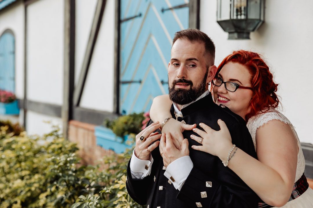Red-haired bride playfully hugging bearded groom in Scottish kilt outdoors.