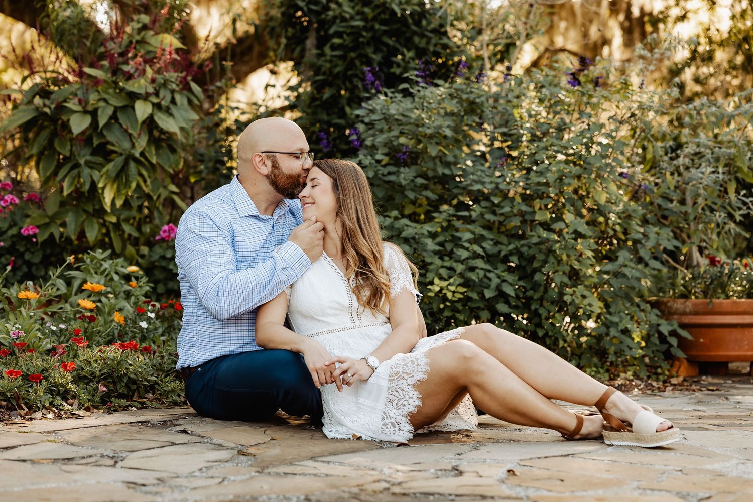 Couple sitting together on stone patio surrounded by colorful garden flowers, sharing a tender moment.