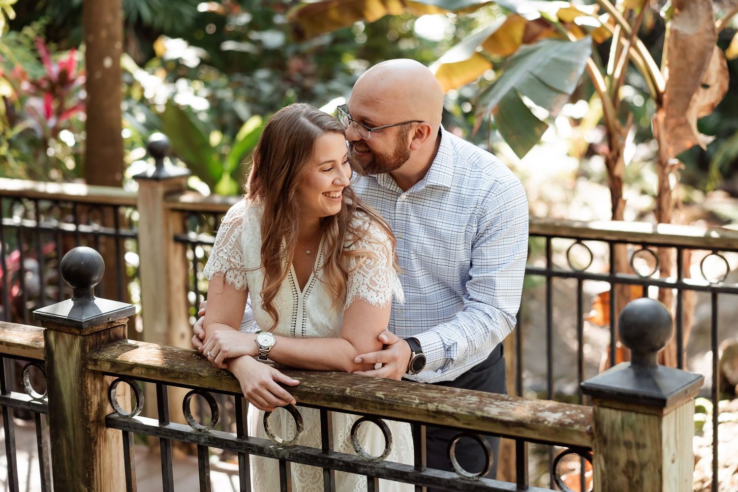 Couple smiling and leaning on iron fence surrounded by lush greenery during outdoor engagement photo session.