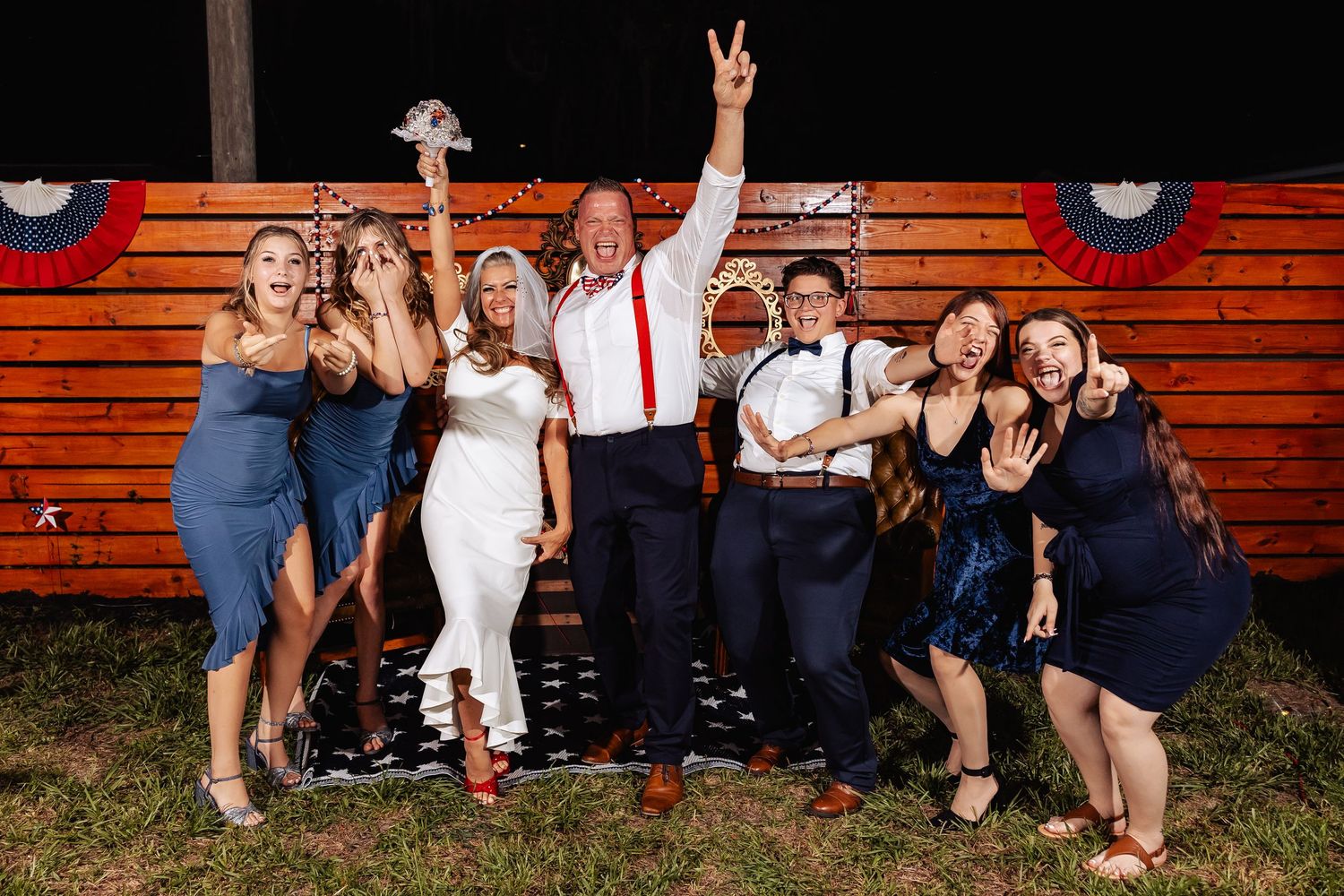 Wedding party celebrating outdoors at night in front of rustic wooden barn with patriotic decorations.