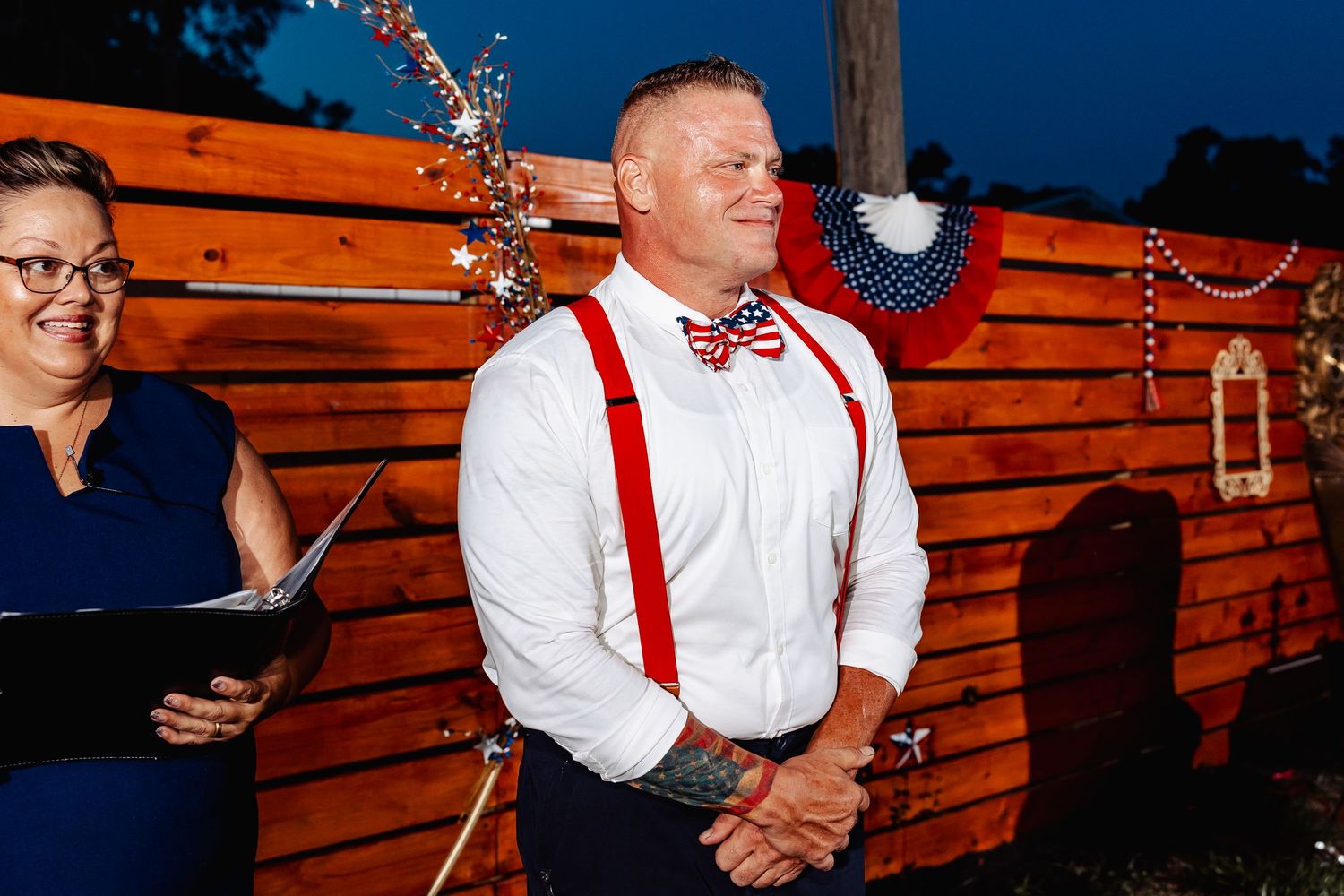 Groom in white shirt with red suspenders and bow tie smiling at outdoor wedding ceremony.