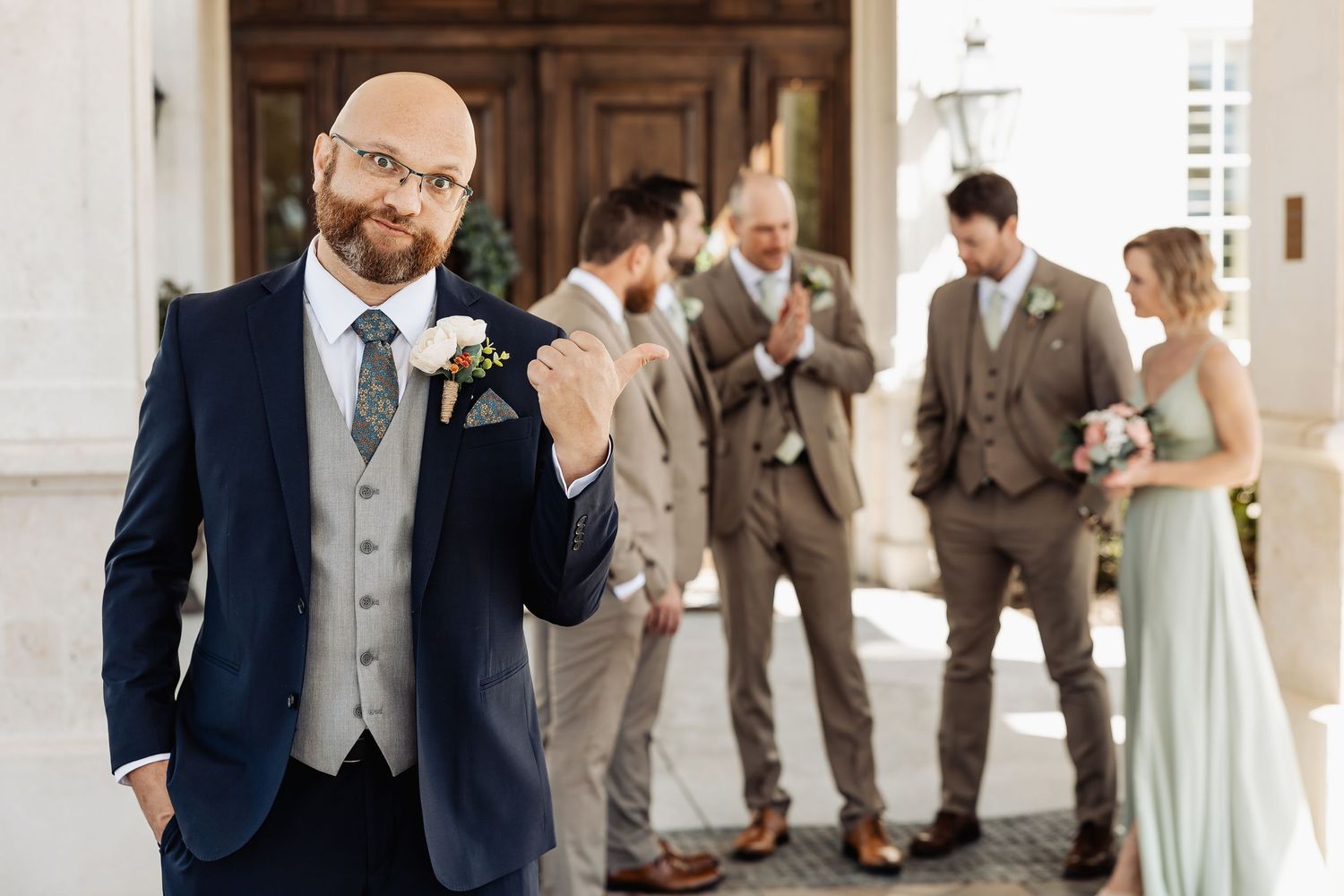 Bald groom in navy suit gives thumbs up while groomsmen in tan suits celebrate behind him outdoors.