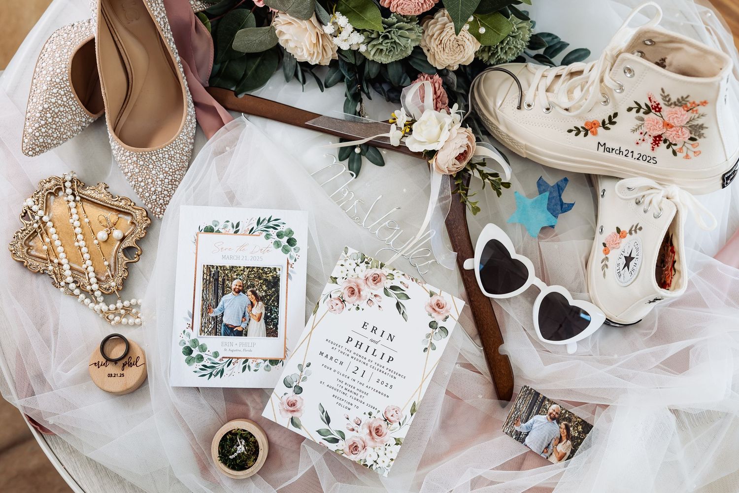 Flat lay of wedding details including floral invitation, pearl jewelry, custom Converse sneakers, and bridal accessories.