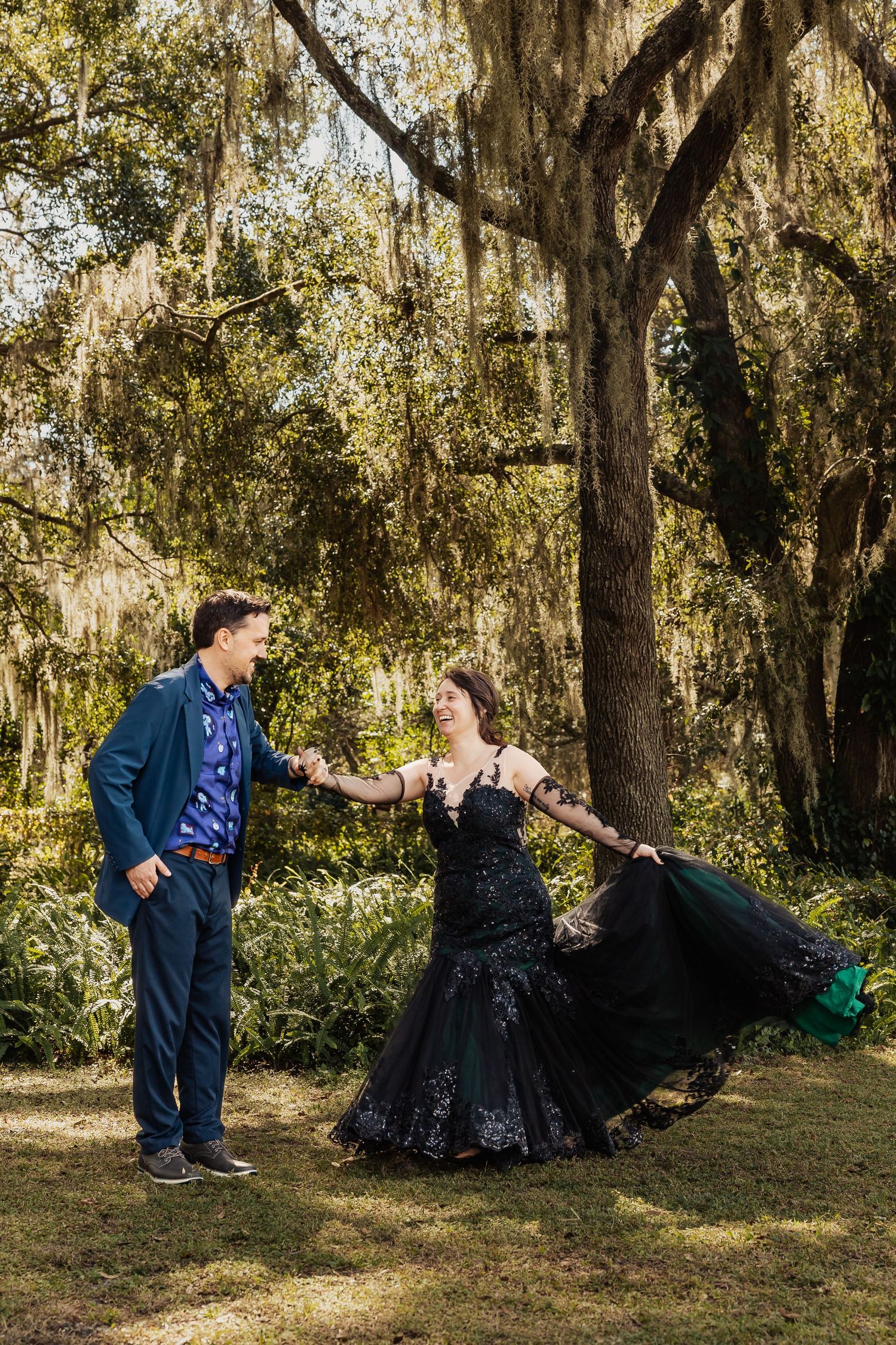 Joyful bride in black mermaid gown twirls with groom under Spanish moss-covered oak trees at outdoor wedding.