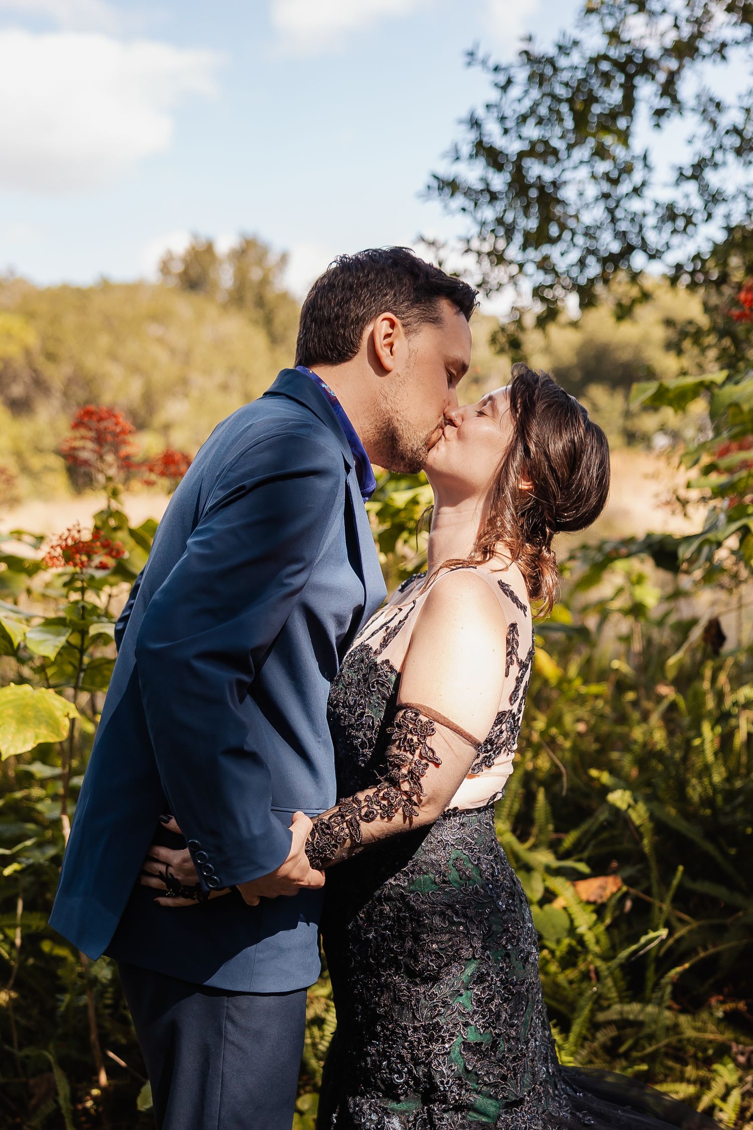 Couple sharing romantic kiss in garden, bride in black lace gown and groom in blue suit.