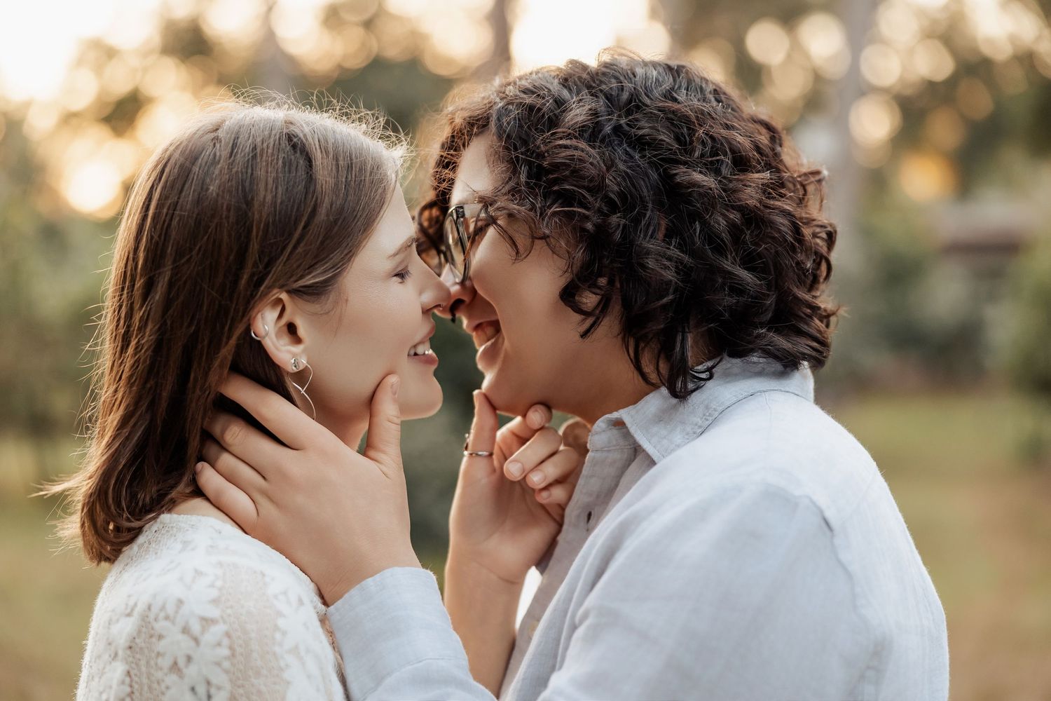 Two women laughing joyfully face to face outdoors in golden sunlight, holding each other gently.