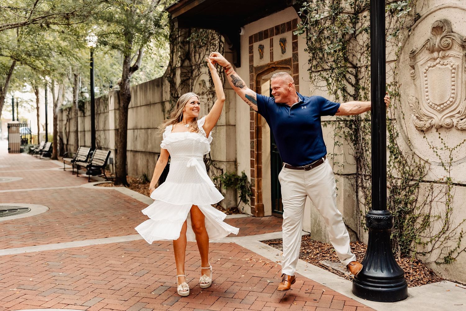 Joyful couple dancing and twirling on a brick walkway surrounded by lush greenery and vintage decor.