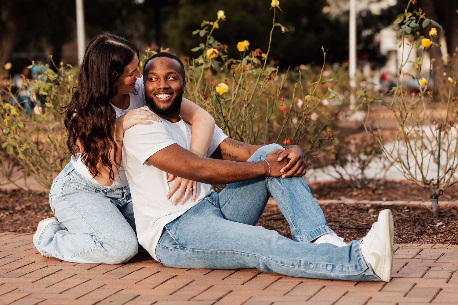 Happy interracial couple sitting together on brick path surrounded by yellow roses in garden.