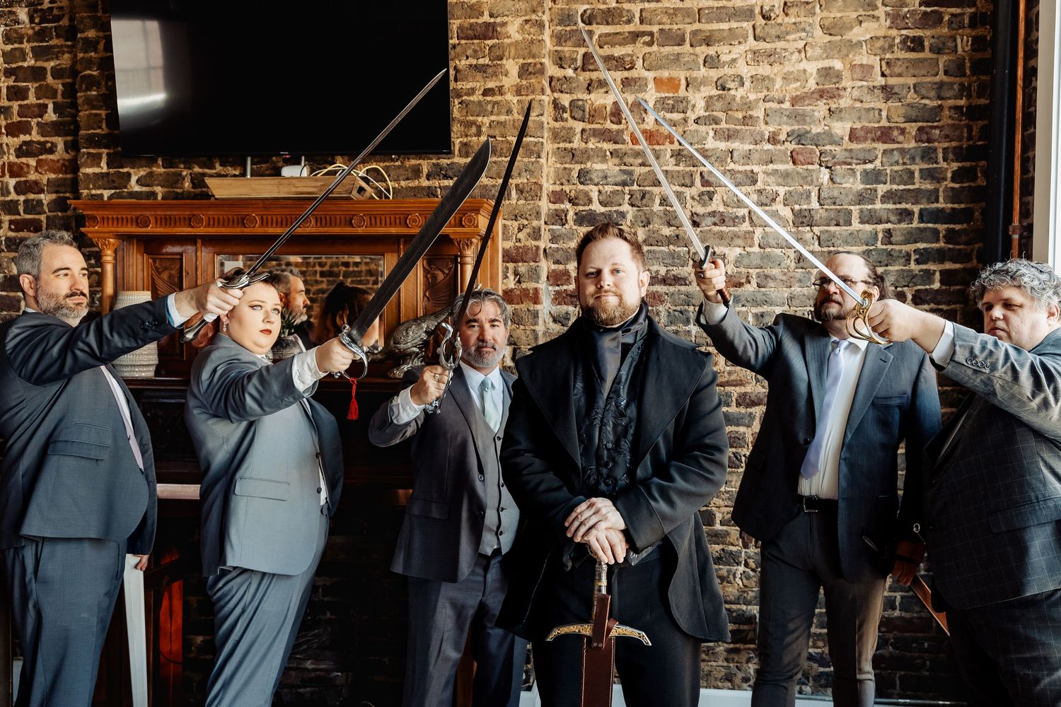 Groom and groomsmen in suits raising swords inside a brick-walled wedding venue.
