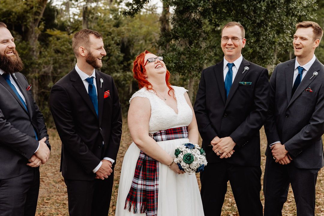 Wedding party in formal attire with kilts stands together outdoors.