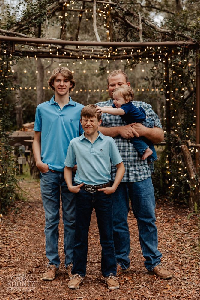 Family portrait of four people in coordinated blue outfits standing under string lights outdoors.