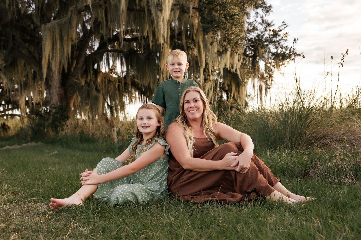 A casual family portrait on grass with Spanish moss trees creating a dreamy backdrop at sunset.
