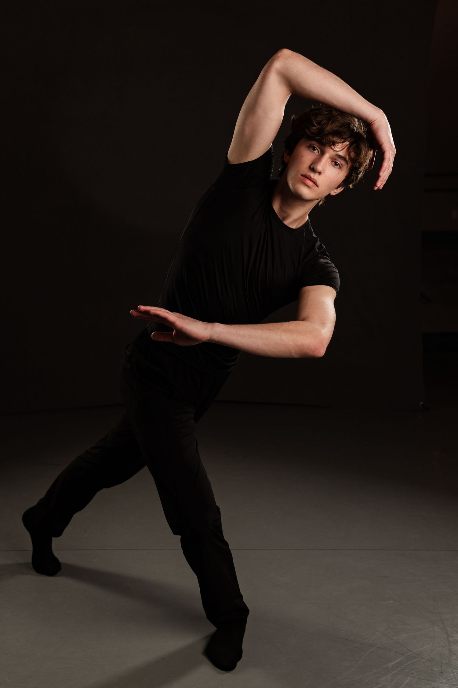 Dancer in black attire performs expressive modern dance pose against dark background.