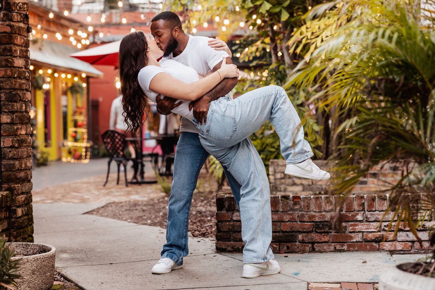 Romantic dip pose captured in front of a rustic brick building with string lights and greenery.