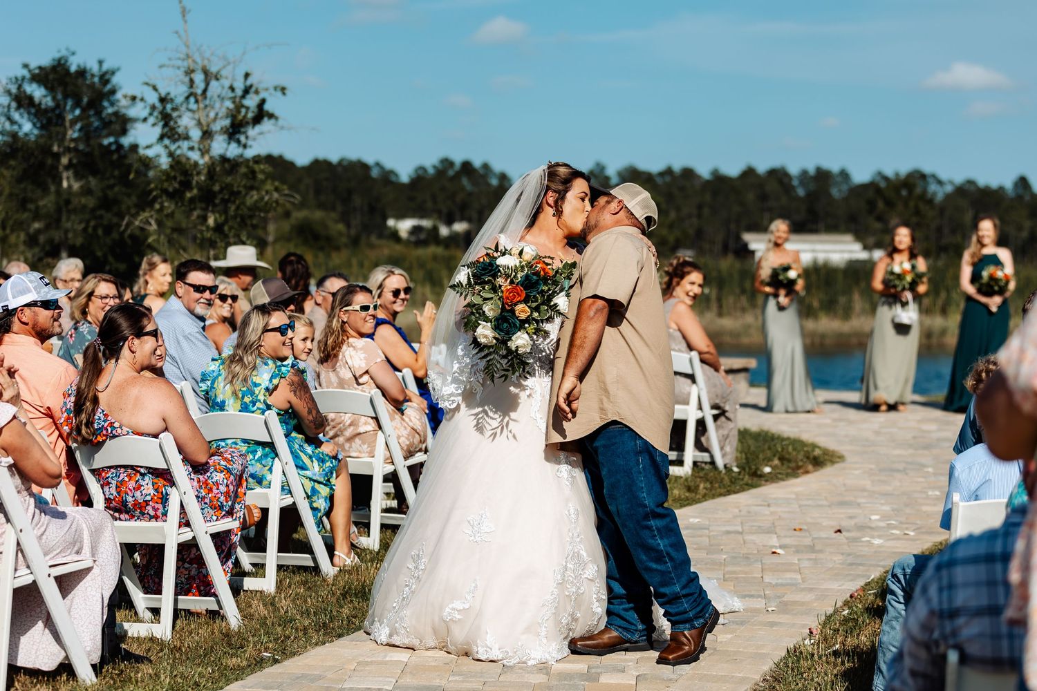 Wedding guests seated in white chairs watch a ceremony on a lakeside path under blue skies.