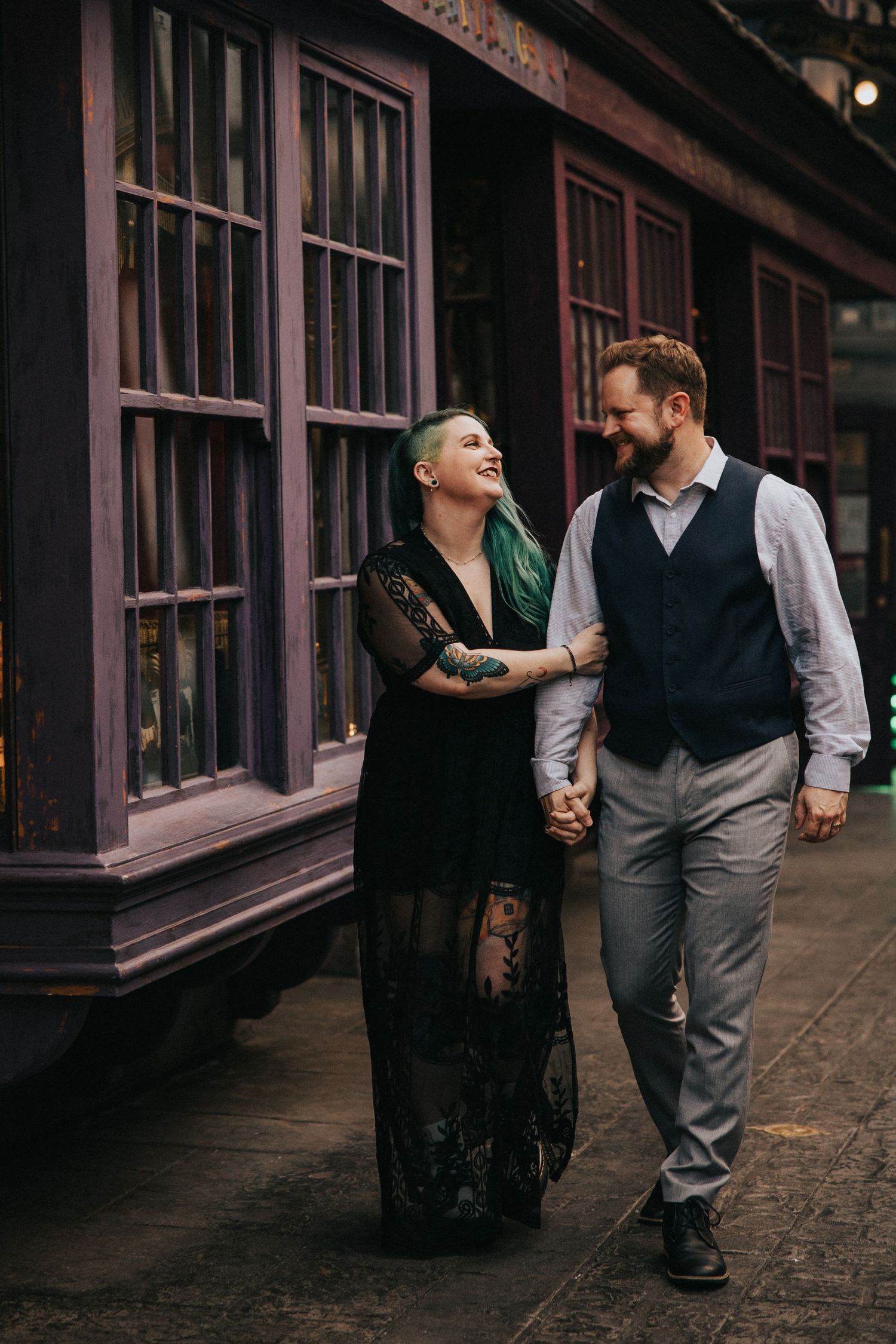 A couple walks together on a historic street with vintage windows in the background.