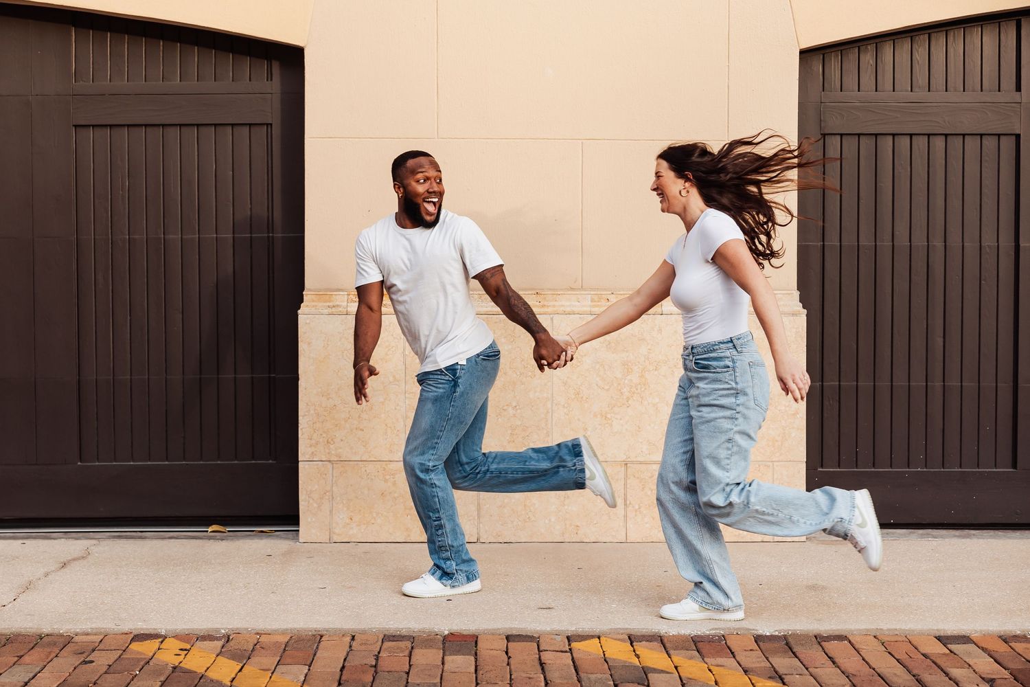 Couple in matching white t-shirts and jeans running playfully in front of dark wooden doors.