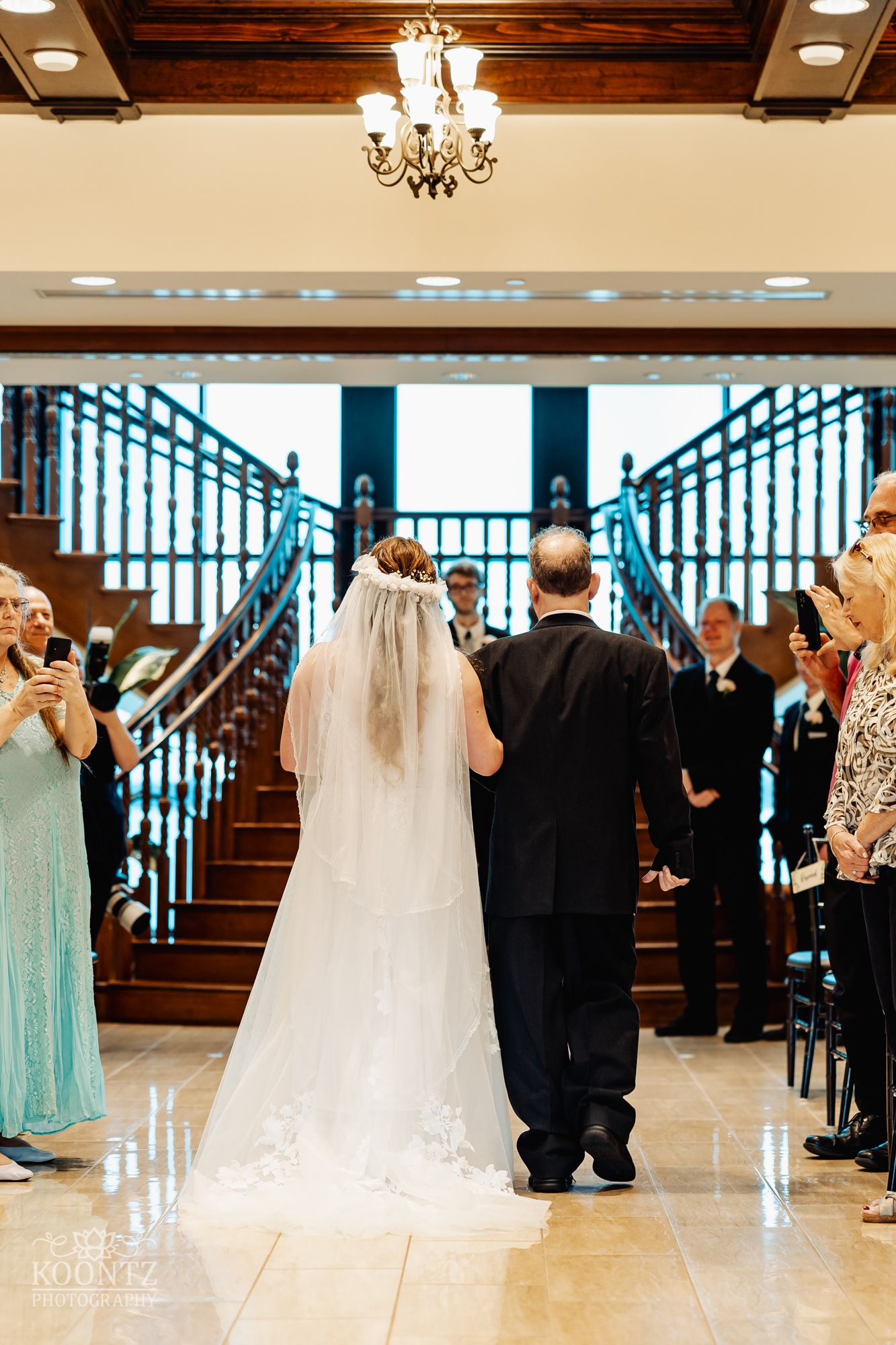 Wedding ceremony in elegant venue with chandelier and blue staircase as couple walks down aisle.