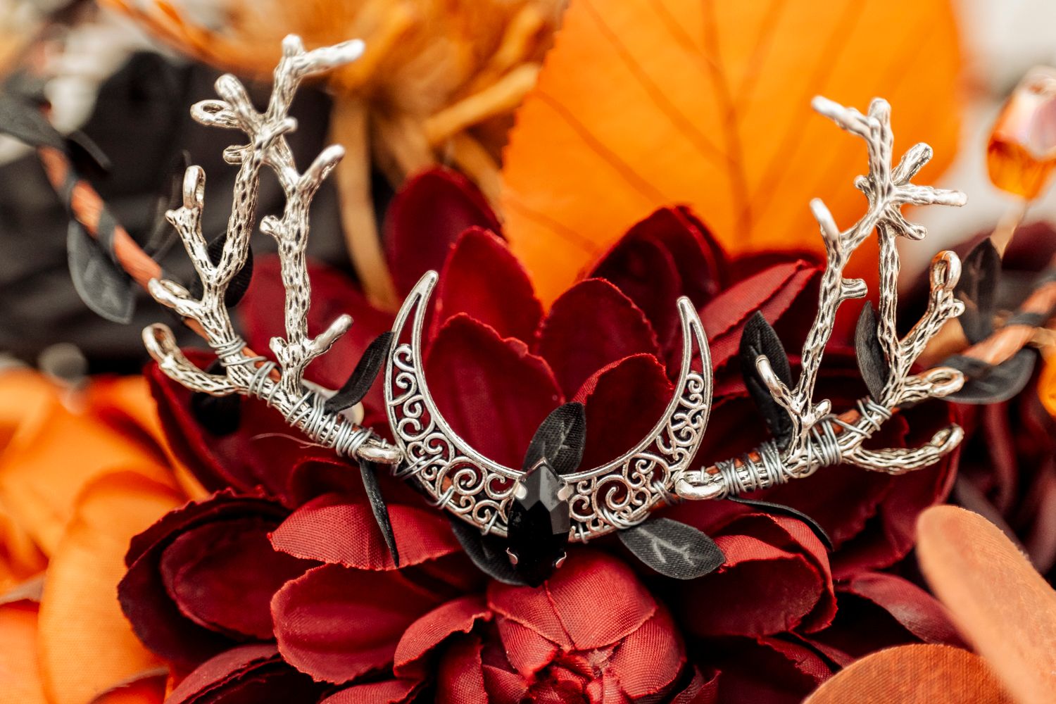 A decorative silver crown with intricate antler designs and crescent moons rests atop deep red flowers against orange leaves.