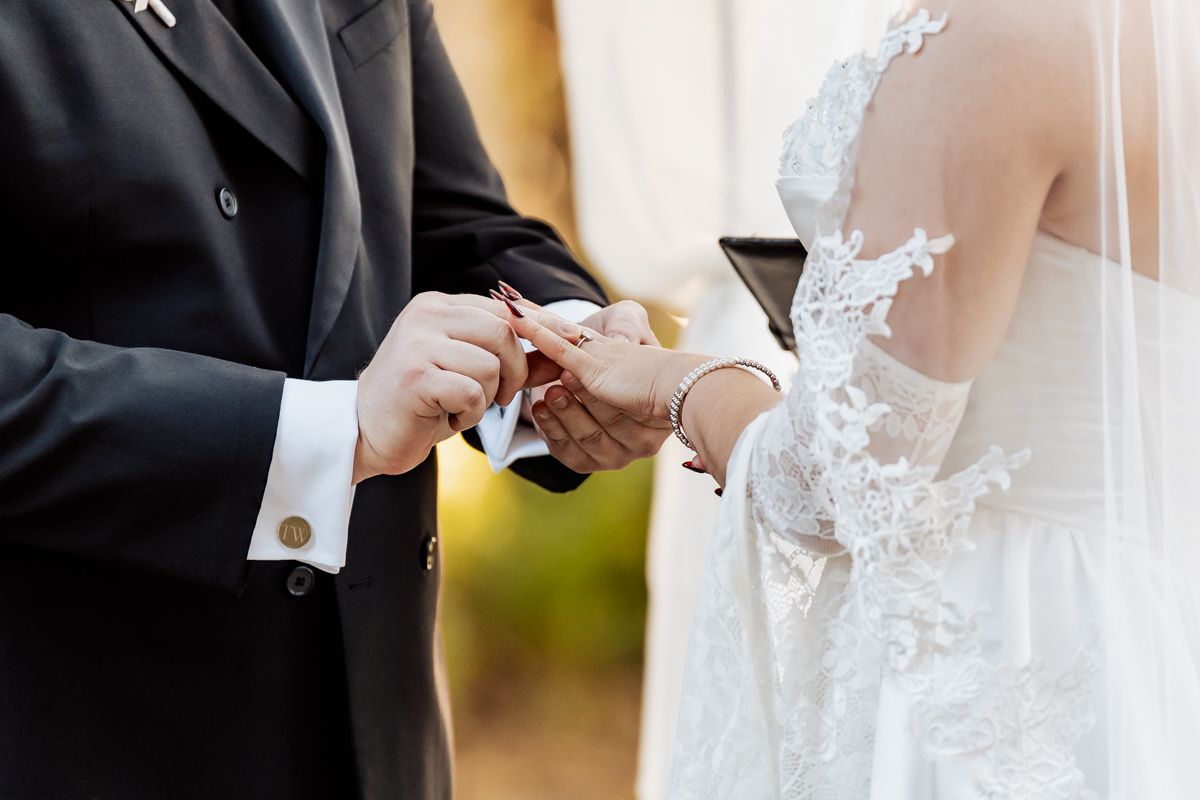 Close-up of wedding ceremony hands exchanging rings with lace dress detail visible.