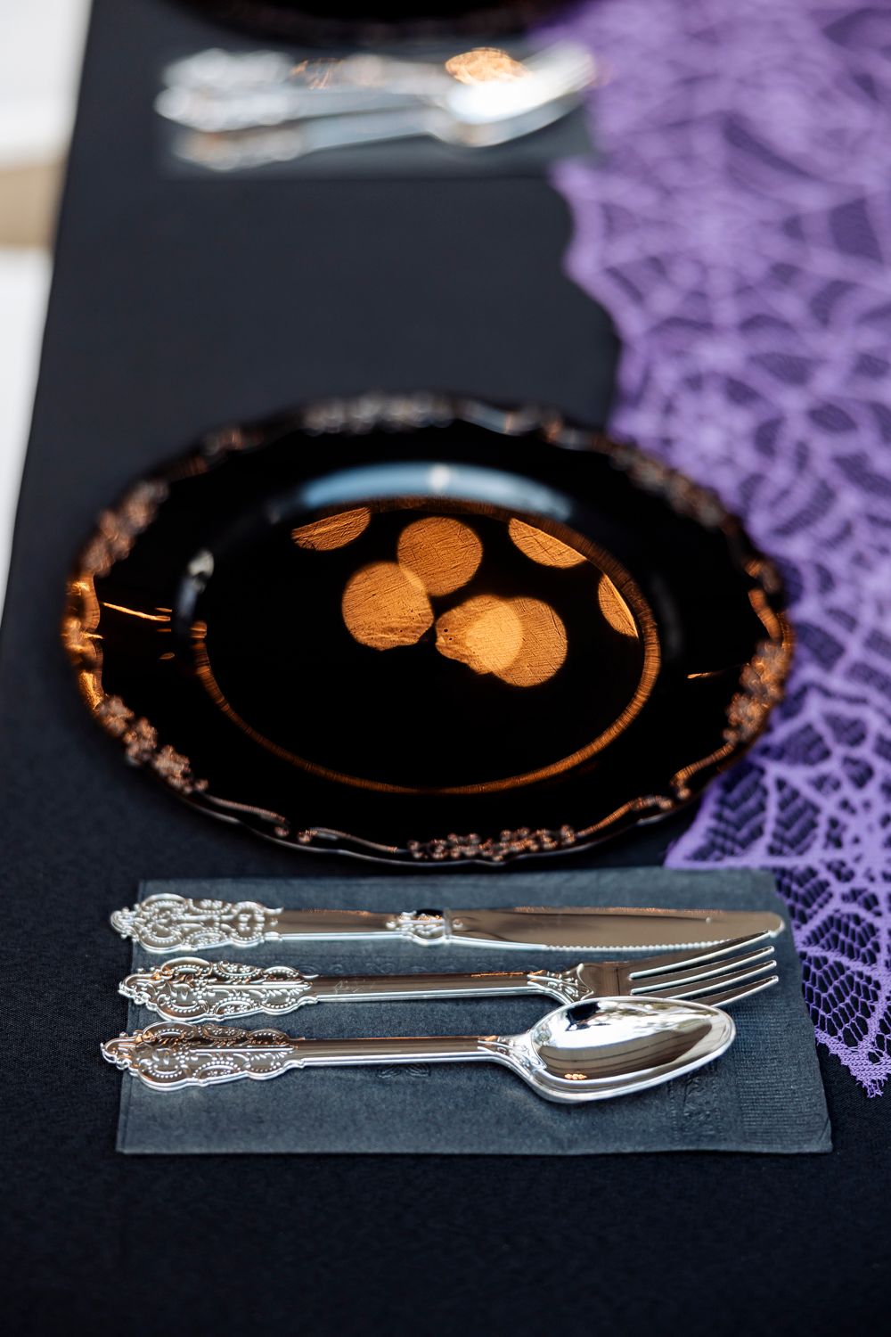 Elegant place setting with black plate, silver utensils and purple lace tablecloth.