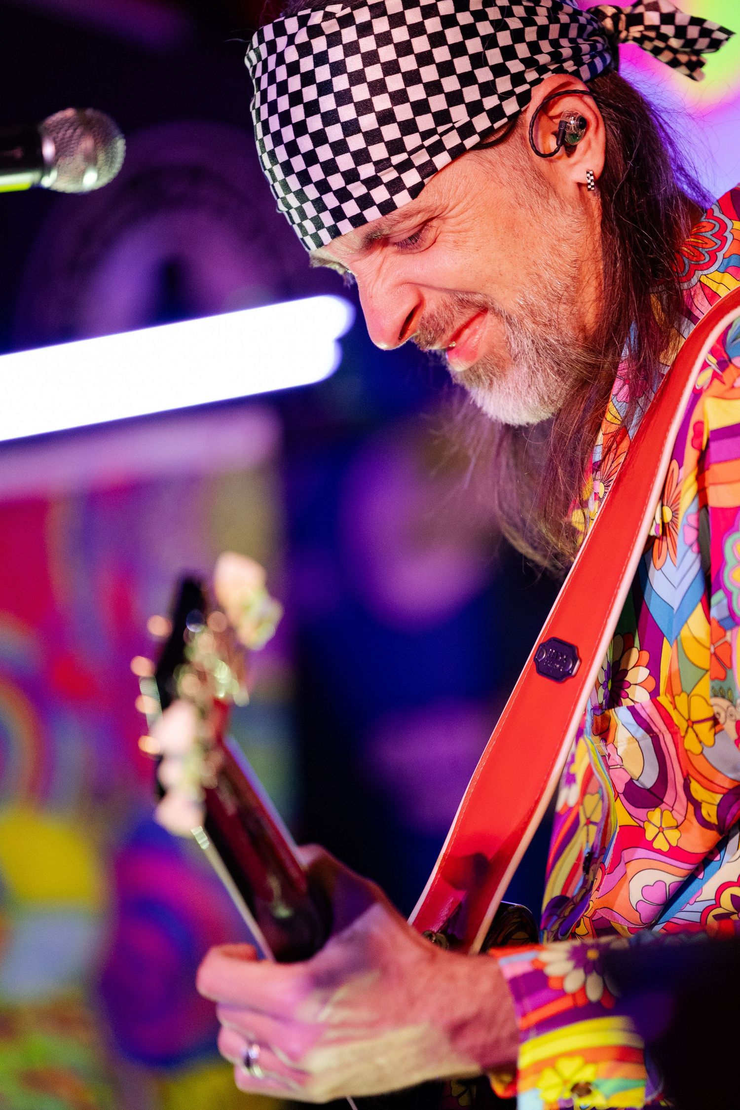 Close-up of older guitarist in psychedelic shirt and checkered headband playing red electric guitar.